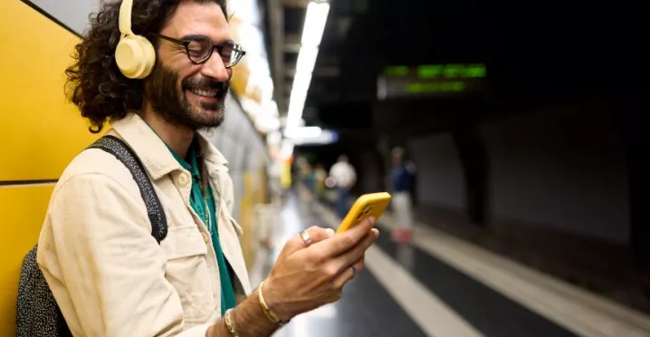 Persoon met een koptelefoon en bril gebruikt een gele smartphone op een treinperron