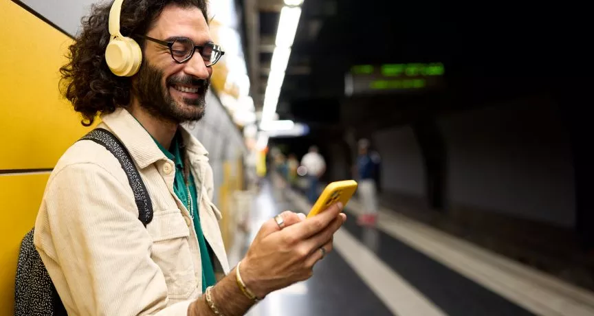 Persoon met een koptelefoon en bril gebruikt een gele smartphone op een treinperron