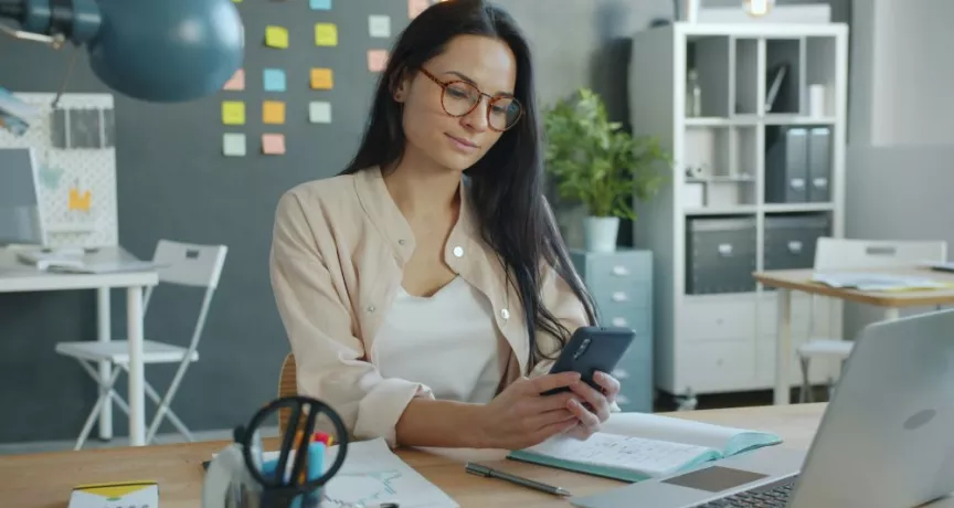 Medewerker gebruikt een smartphone aan een bureau met een laptop en financiële documenten.