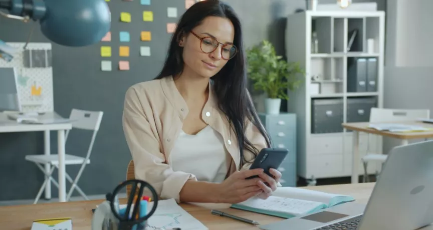 Une personne utilise un smartphone à un bureau avec un ordinateur portable et des documents financiers.