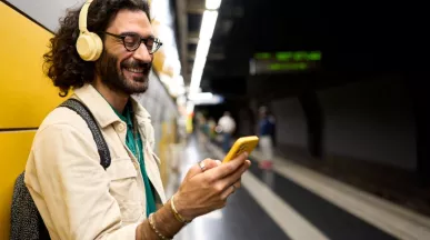 Persoon met een koptelefoon en bril gebruikt een gele smartphone op een treinperron